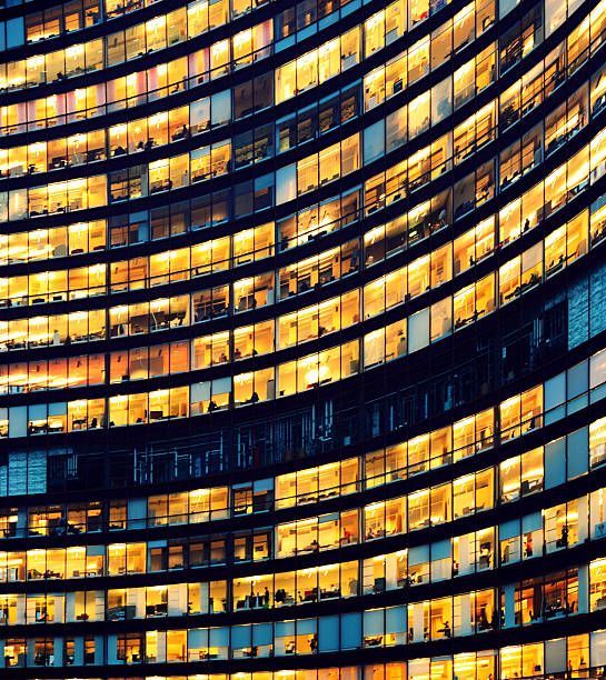 The image depicts workers working late in an office at night.  The employees are seen as silhouettes against their lit offices that have large windows.  The building is lit by the "blue hour" evening sky.  The building and its windows cover the entire canvas of the image.  It is apparent that the building may have some sort of curvature to the architecture.  While most of the building reflects patterns of yellow light, a small portion of it reflects the blue light.  There is no border around the image.  The windows on the edges and corners of the image are truncated in the view.